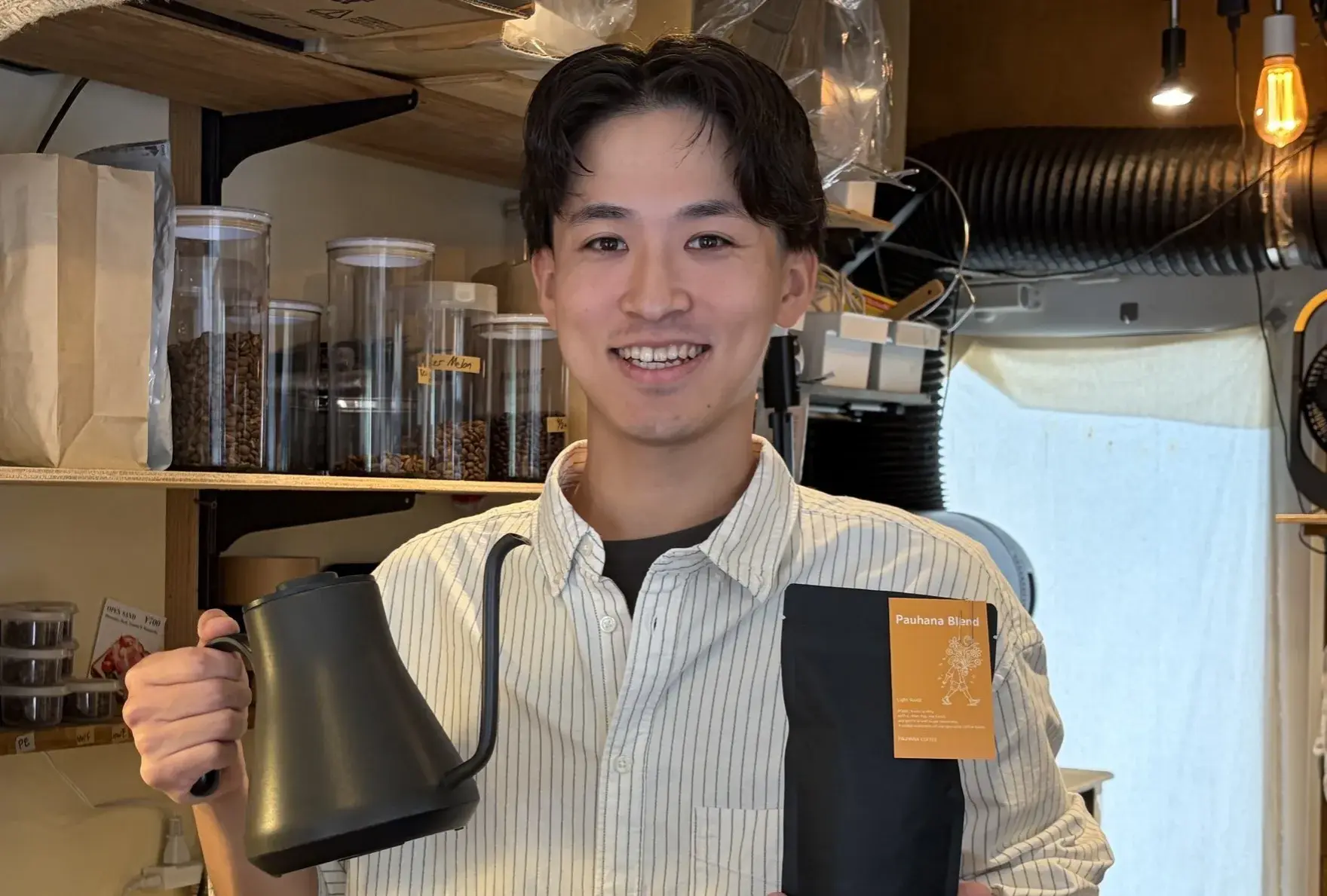 コーヒー豆の瓶が並ぶ店内で、ケトルとPauhana Blendの袋を手にする吉岡仁さん